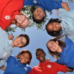 A diverse group of students standing in a circle outdoors, smiling and looking down toward the camera.